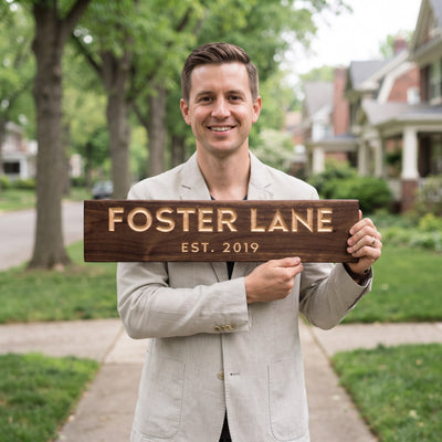 A smiling man in a light jacket standing on his front walkway on a quiet tree-lined street holds a personalized wood sign that reads Foster Lane