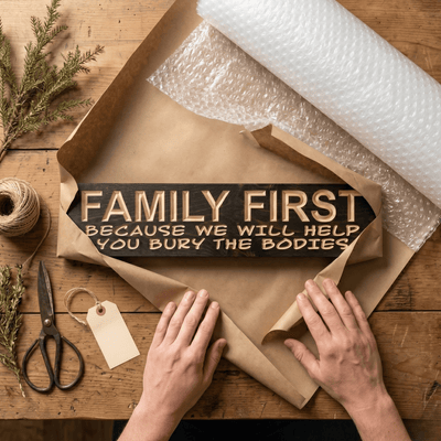 Family First funny farmhouse sign being hand-wrapped in kraft paper and bubble wrap on a rustic workshop table with twine, scissors, and blank gift tag
