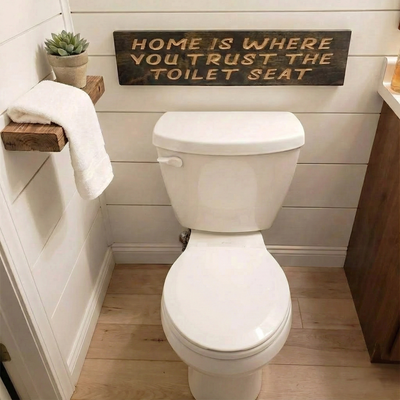 "Home Is Where You Trust The Toilet Seat" wooden sign hanging on a white shiplap wall above a standard toilet in a farmhouse style bathroom.