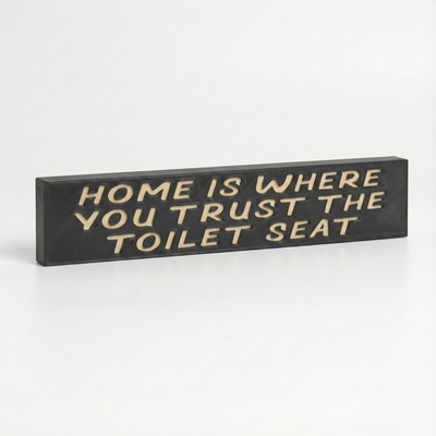 Angled studio view of a rustic wood sign reading "Home Is Where You Trust The Toilet Seat" featuring deep V-carved lettering and dark stain.