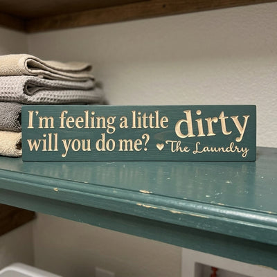 A teal Laundry Room Sign with "I'm feeling a little dirty will you do me? The Laundry" sits on a shelf with folded towels from Bluegrass Gifts