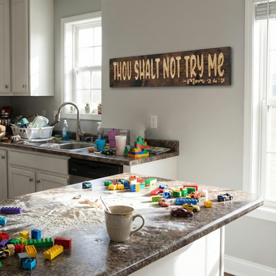 The "Thou Shalt Not Try Me - Mom 24:7" wooden sign hanging on a wall in a real-life messy kitchen setting with toys on the counter.