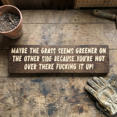A Garden Shed Sign made of dark wood with light carved letters sits on a rustic wooden table next to a potted plant and gardening gloves from Bluegrass Gifts