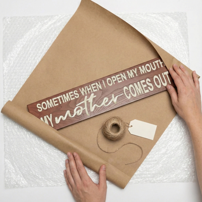 Overhead view of a funny carved wood sign being wrapped in brown craft paper with twine and a gift tag.
