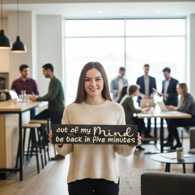 A smiling professional woman holding the "Out of My Mind" wood sign in a modern open-concept office setting.