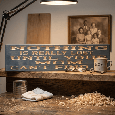 Funny mom wood sign on a workbench with fresh wood shavings, a MOM coffee mug, vintage family portrait, and workshop lamp — handcrafted in Rineyville, Kentucky.