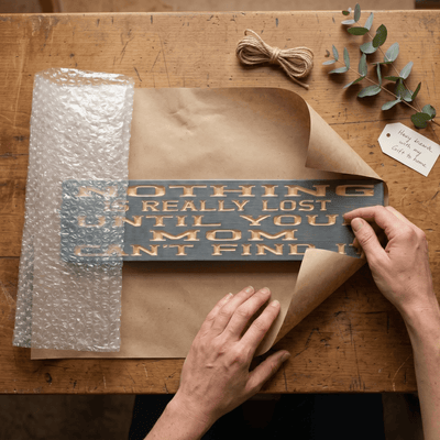 Funny mom wood sign being carefully wrapped in kraft paper with bubble wrap, twine, and a handwritten gift tag — handmade gift packaging from Bluegrass Gifts.