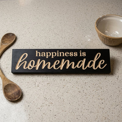 A black Happiness Is Homemade Sign rests on a light speckled kitchen counter next to wooden spoons and a bowl from Bluegrass Gifts