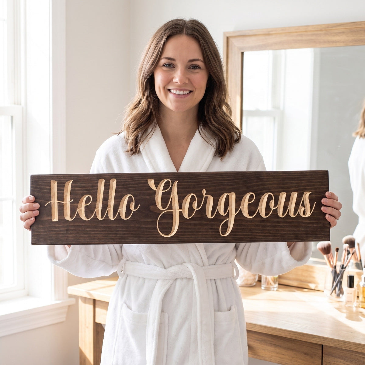 A smiling woman in a white robe holds the Hello Gorgeous sign by her vanity