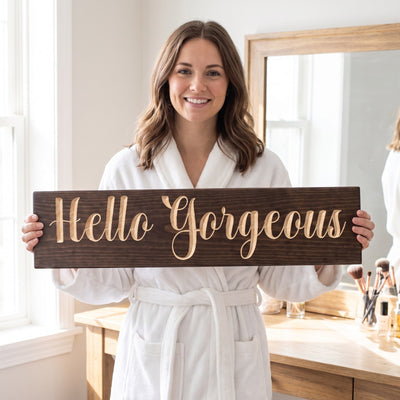 A smiling woman in a white robe holds the Hello Gorgeous sign by her vanity