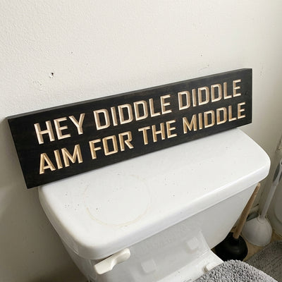 A black Hey Diddle Diddle Aim for the Middle Sign rests on a white toilet tank, a plunger visible nearby from Bluegrass Gifts