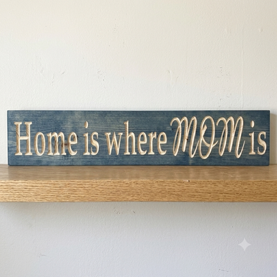 A straight-on view of the blue "Home Is Where Mom Is Sign" with light carved letters, resting on a wooden shelf from Bluegrass Gifts