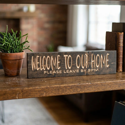 A dark Homebody Sign with carved light letters rests on a wooden shelf beside a green plant and old books, from Bluegrass Gifts