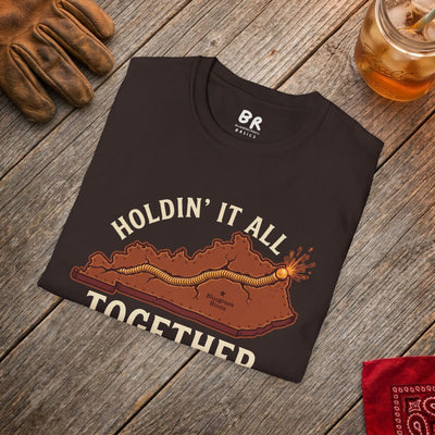 The dark brown Kentucky Shirt - Holdin' It All Together tee is folded on a wooden surface with work gloves and a drink