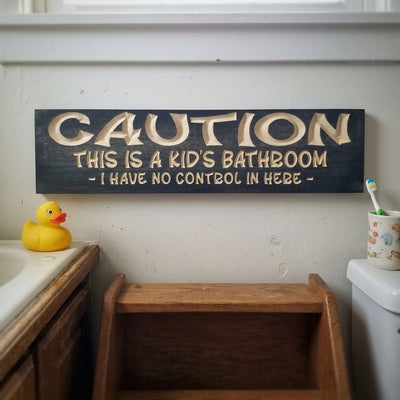 A dark Kids Bathroom Sign with light carved letters hangs on a white wall above a sink with a rubber duck, from Bluegrass Gifts