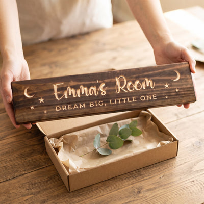 Hands holding a personalized wood sign that reads Emma's Room over a kraft gift box with tissue paper and twine