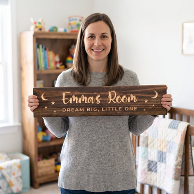 A smiling mom in a soft sweater standing in a cheerful child's bedroom holds a personalized wood sign that reads Emma's Room