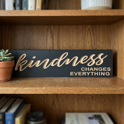 A Kindness Matters Sign sits on a wooden bookshelf next to a small green plant in a terracotta pot, from Bluegrass Gifts