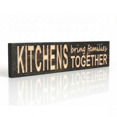 A side-view of a handcrafted dark wood sign by Bluegrass Gifts with CNC-carved lettering that reads "KITCHENS bring families TOGETHER" on a reflective white surface.