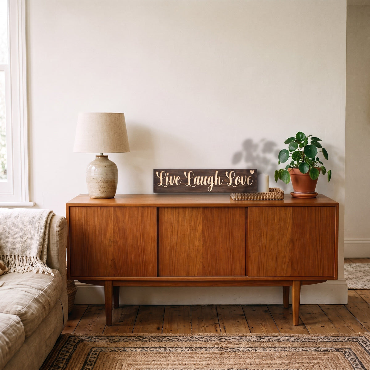 The Live Laugh Love sign leaning on a wooden sideboard in a living room beside a table lamp and a potted plant