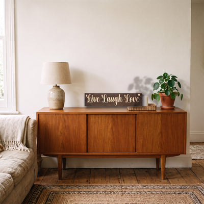 The Live Laugh Love sign leaning on a wooden sideboard in a living room beside a table lamp and a potted plant