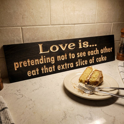 A dark wood sign, "Love Is Pretending Not to See Each Other Eat That Extra Slice of Cake," rests on a kitchen counter with a cake slice, from Bluegrass Gifts