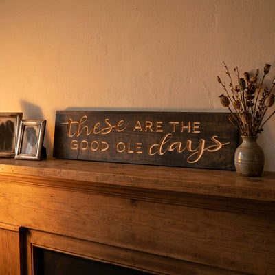 Dark wood Mantel Sign with carved light letters on a wooden mantelpiece, next to silver frames and dried flowers, from Bluegrass Gifts