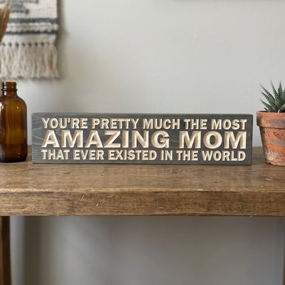 A dark gray carved wood sign with light text, "YOU'RE PRETTY MUCH THE MOST AMAZING MOM THAT EVER EXISTED IN THE WORLD," sits on a rustic wooden table with a brown bottle and a small plant, providing Mom Decor from Bluegrass Gifts