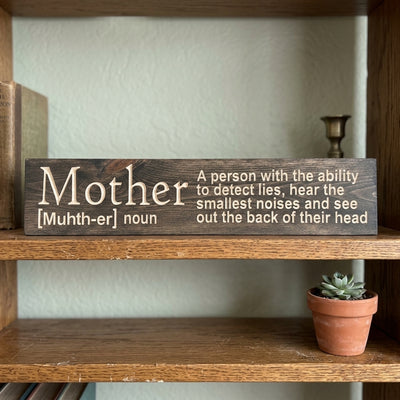 A dark wood Mom Sign with the "Mother" definition sits on a wooden shelf next to old books and a small succulent from Bluegrass Gifts