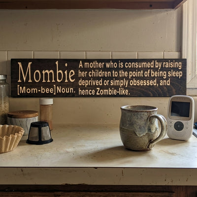 The Mombie Sign rests on a kitchen counter next to a coffee mug and baby monitor, from Bluegrass Gifts