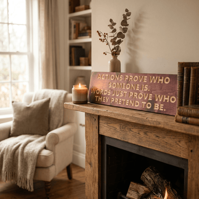 Motivational wood sign reading "Actions Prove Who Someone Is" displayed on a rustic reclaimed wood mantle beside a candle, dried eucalyptus, and leather-bound books — rustic farmhouse living room wall decor.