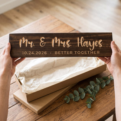 Hands holding a personalized wood sign that reads Mr. & Mrs. Hayes over a kraft gift box with tissue paper and twine