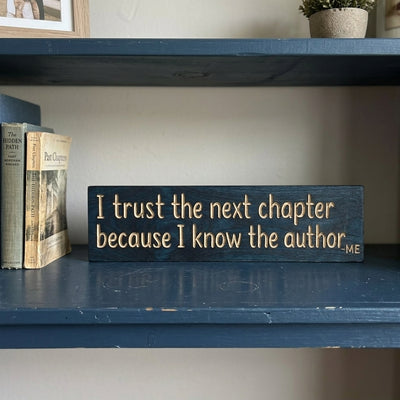 A close-up of the dark blue New Beginnings Sign with bare-wood carved letters resting on a shelf next to old books from Bluegrass Gifts