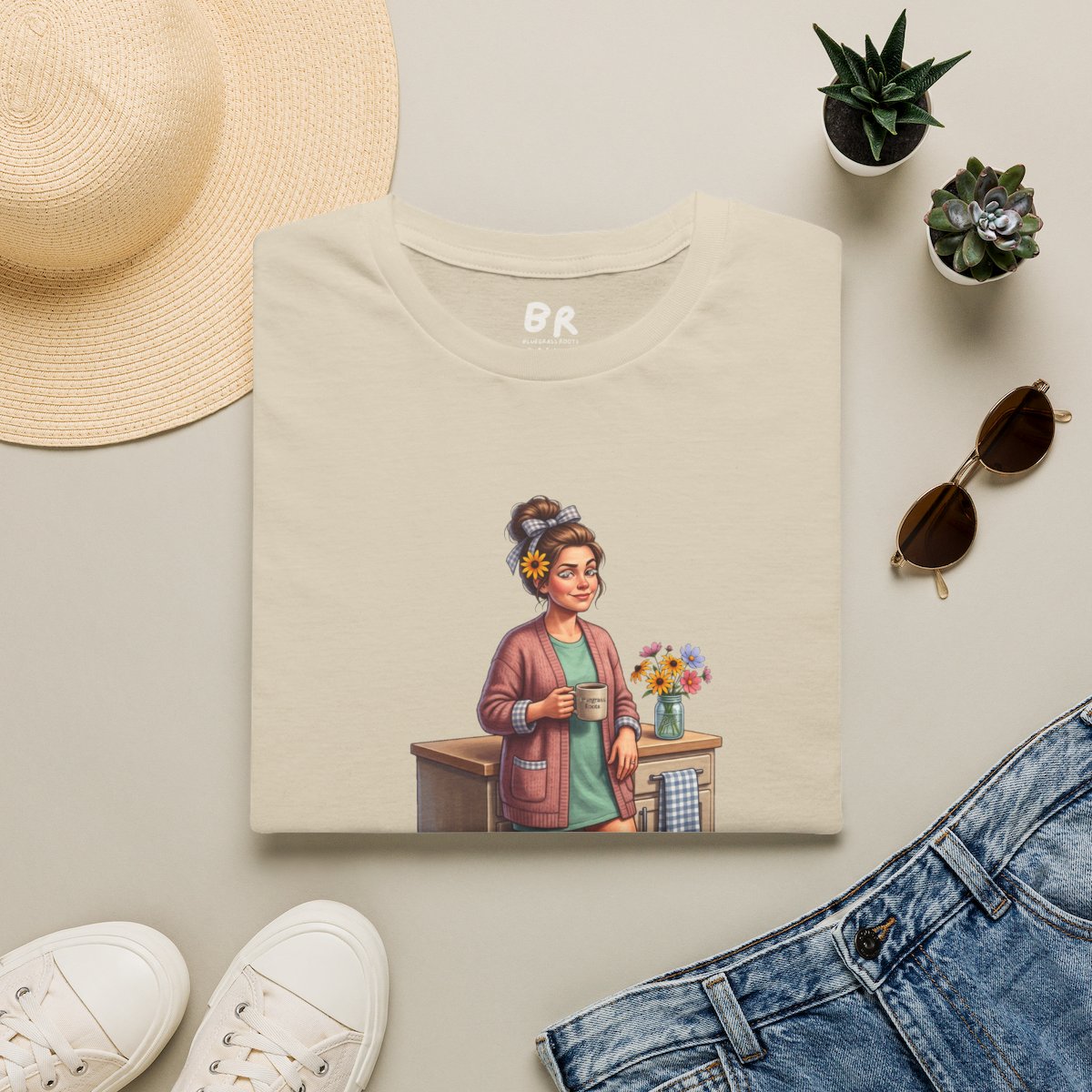 A folded beige Overstimulated Shirt - Overstimulated Moms Club with the character graphic, surrounded by accessories on a flat surface