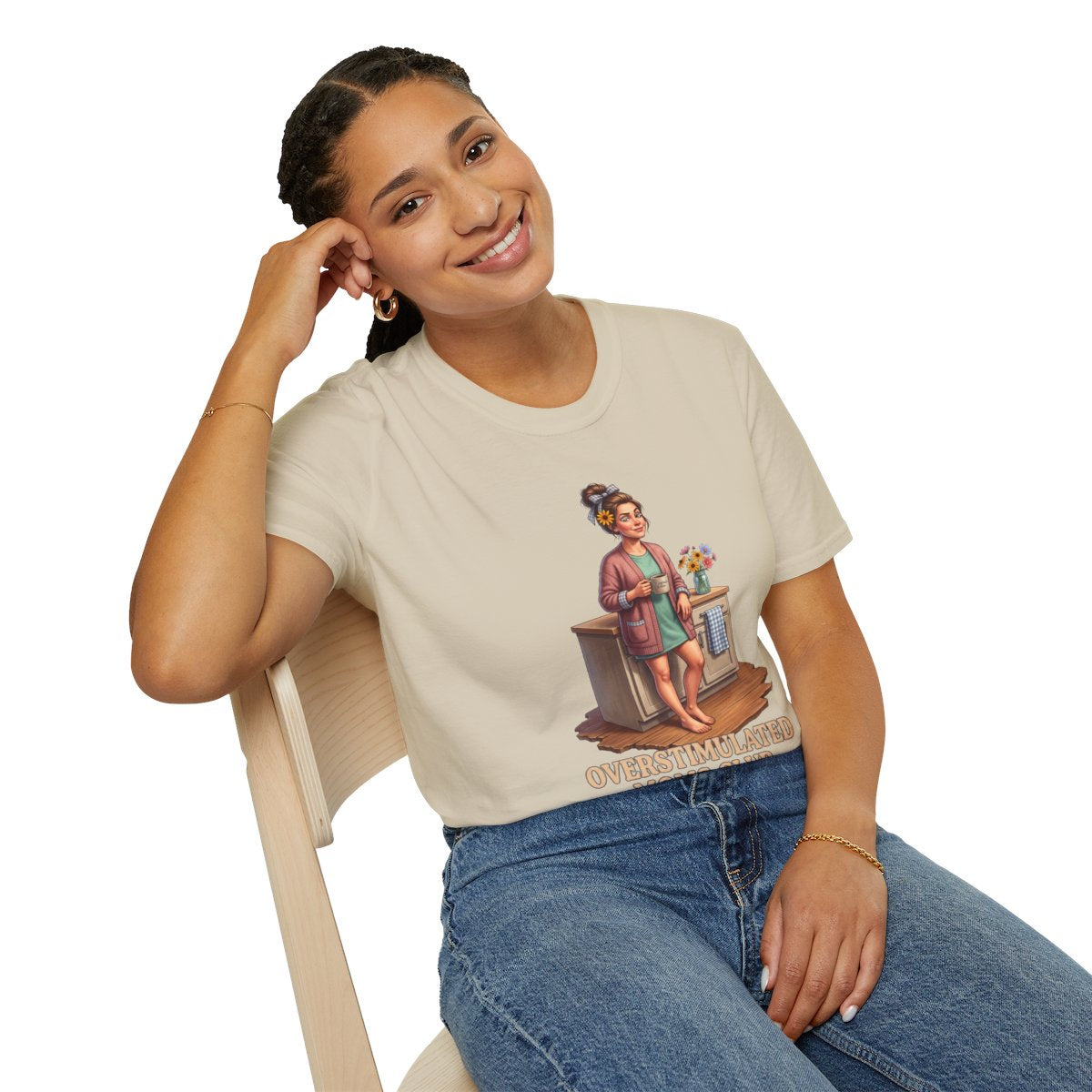 A woman sitting on a light wooden chair, smiling, wearing a beige Overstimulated Shirt - Overstimulated Moms Club