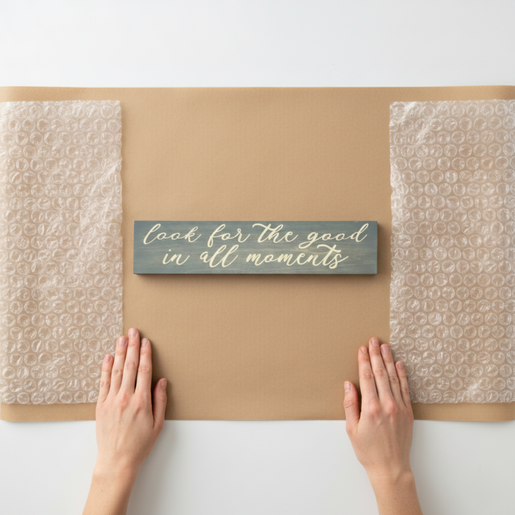 Hands preparing to ship a "Look for the good in all moments" sign, emphasizing the handmade quality and care at Bluegrass Gifts.