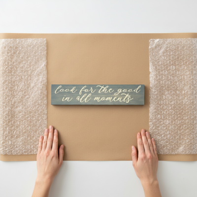 Hands preparing to ship a "Look for the good in all moments" sign, emphasizing the handmade quality and care at Bluegrass Gifts.