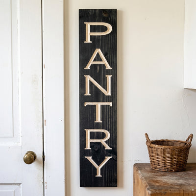 A black vertical Pantry Door Sign with carved light-colored letters hangs on a white door, from Bluegrass Gifts
