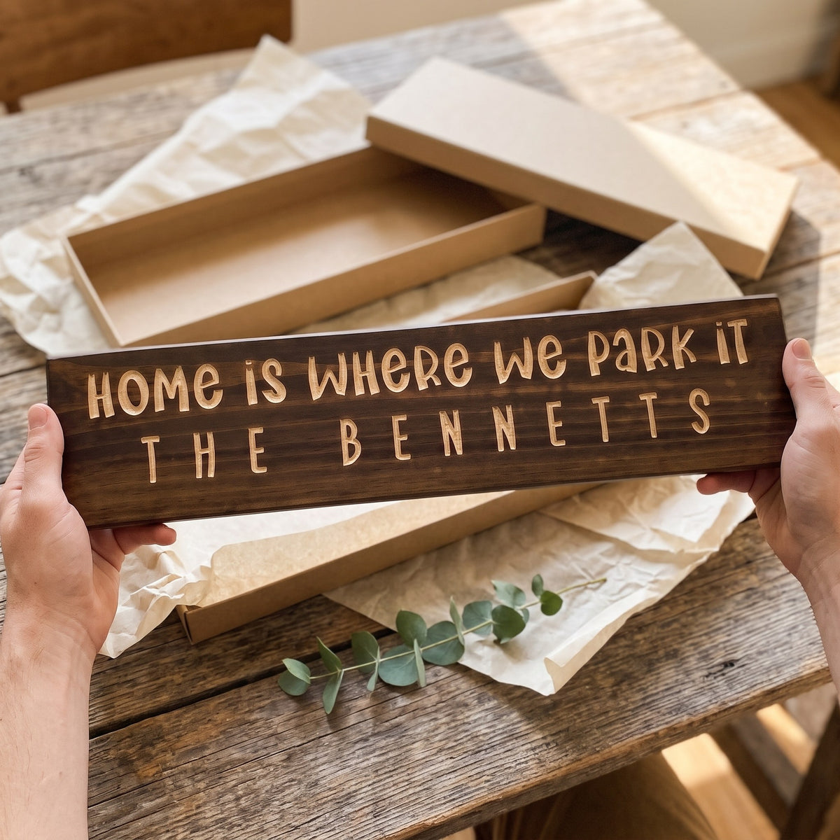 Hands holding a personalized wood sign that reads Home Is Where We Park It over a kraft gift box with tissue paper and twine