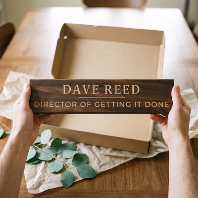 Hands holding a personalized wood sign that reads Dave Reed over a kraft gift box with tissue paper and twine
