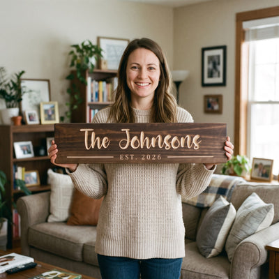 A smiling woman in her thirties in a cozy knit sweater standing in her warm living room holds a personalized wood sign that reads The Johnsons