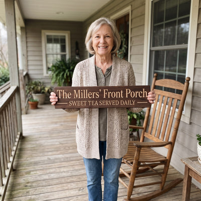 A smiling older woman in a cardigan standing on her front porch beside a wooden rocking chair holds a personalized wood sign that reads The Millers' Front Porch