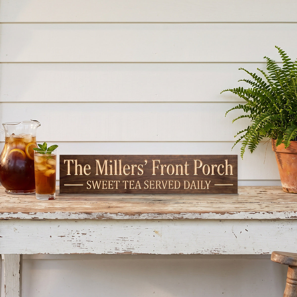 The Millers' Front Porch personalized wood sign standing on a white porch table beside a pitcher of sweet tea and a potted fern