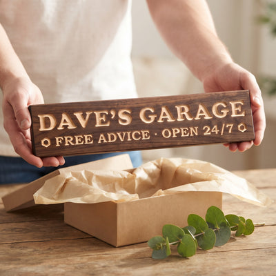 Hands holding a personalized wood sign that reads Dave's Garage over a kraft gift box with tissue paper and twine