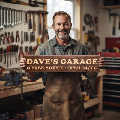 A smiling man in work clothes standing in his tidy garage workshop holds a personalized wood sign that reads Dave's Garage