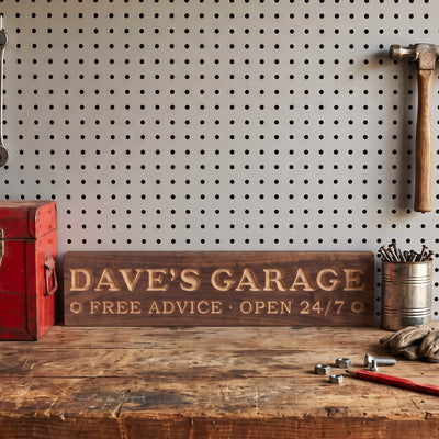 Dave's Garage personalized wood sign standing on a workbench in front of a pegboard, beside a red toolbox, a can of screws and a hammer