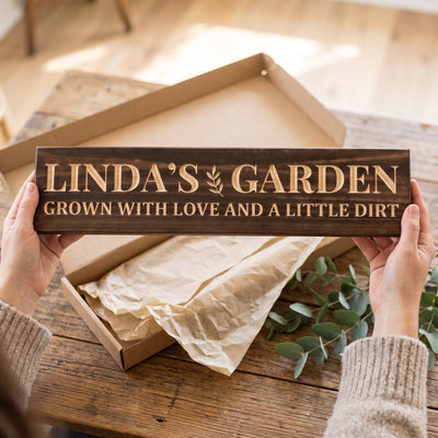 Hands holding a personalized wood sign that reads Linda's Garden over a kraft gift box with tissue paper and twine
