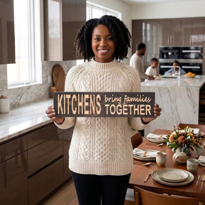 A smiling woman in a modern kitchen holding the "KITCHENS bring families TOGETHER" wood sign, with her family gathered in the background.