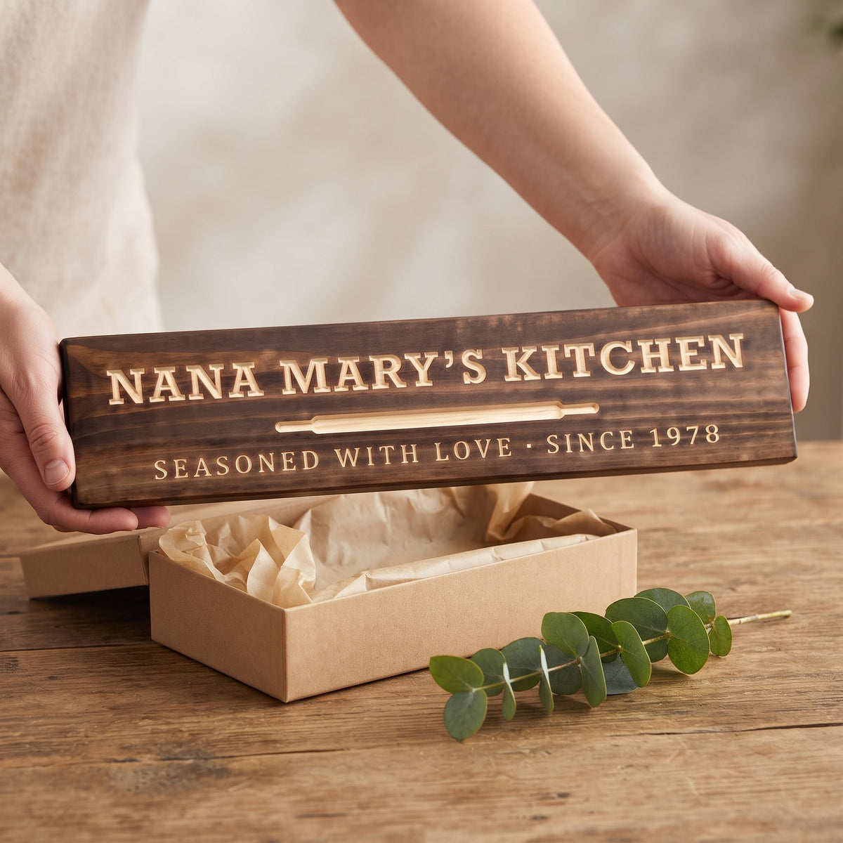 Hands holding a personalized wood sign that reads Nana Mary's Kitchen over a kraft gift box with tissue paper and twine