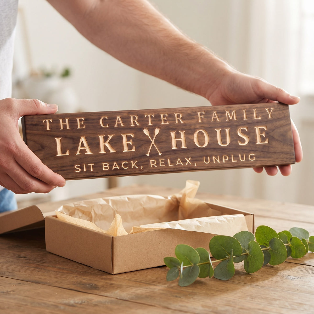 Hands holding a personalized wood sign that reads The Carter Family Lake House over a kraft gift box with tissue paper and twine